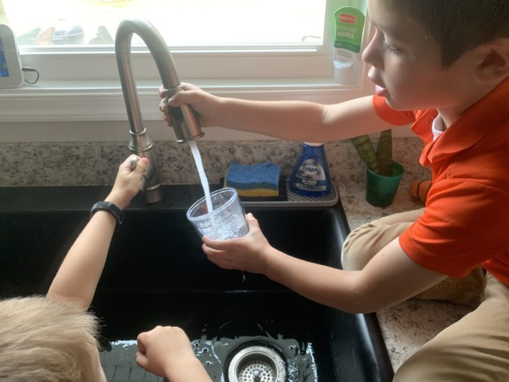 little boy in an orange shirt testing the tap water from the kitchen sink